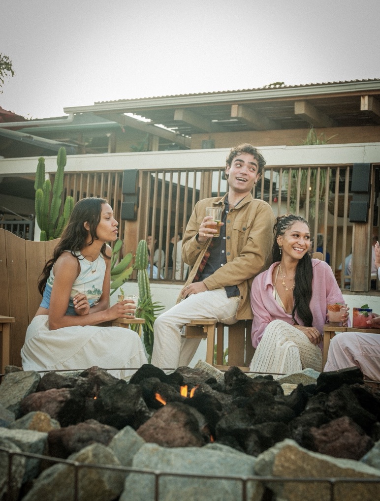 Maria Williams in a pink cardigan laughing with friends around a fire pit, cocktail in hand, for Hedonistas De La Fe
