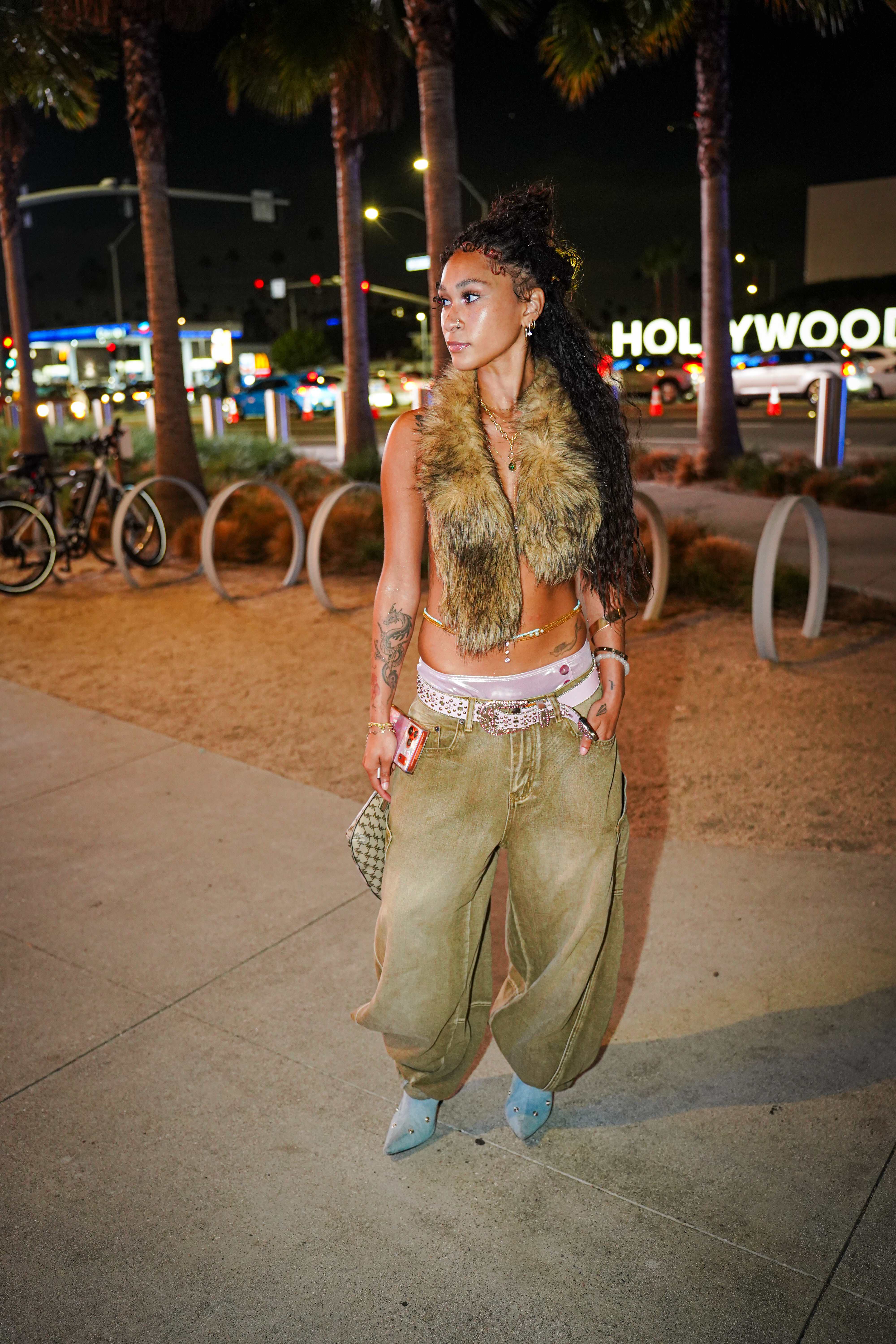 Maria Williams on a Hollywood sidewalk at night in a faux fur vest, olive baggy jeans, a pink studded belt, and denim boots, looking to the side