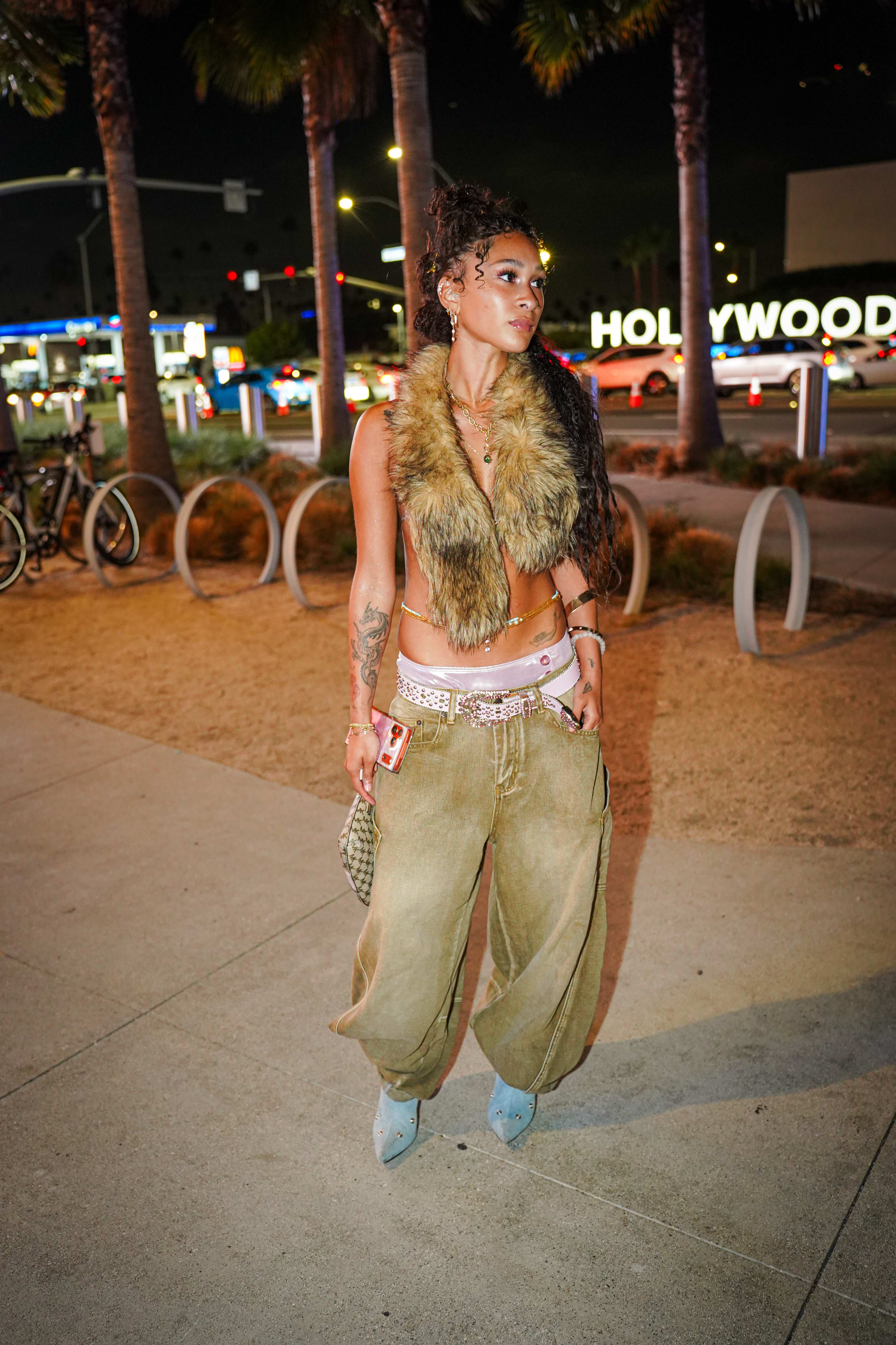 Maria Williams standing under palm trees with the lit Hollywood sign behind her, wearing a faux fur vest and olive baggy jeans