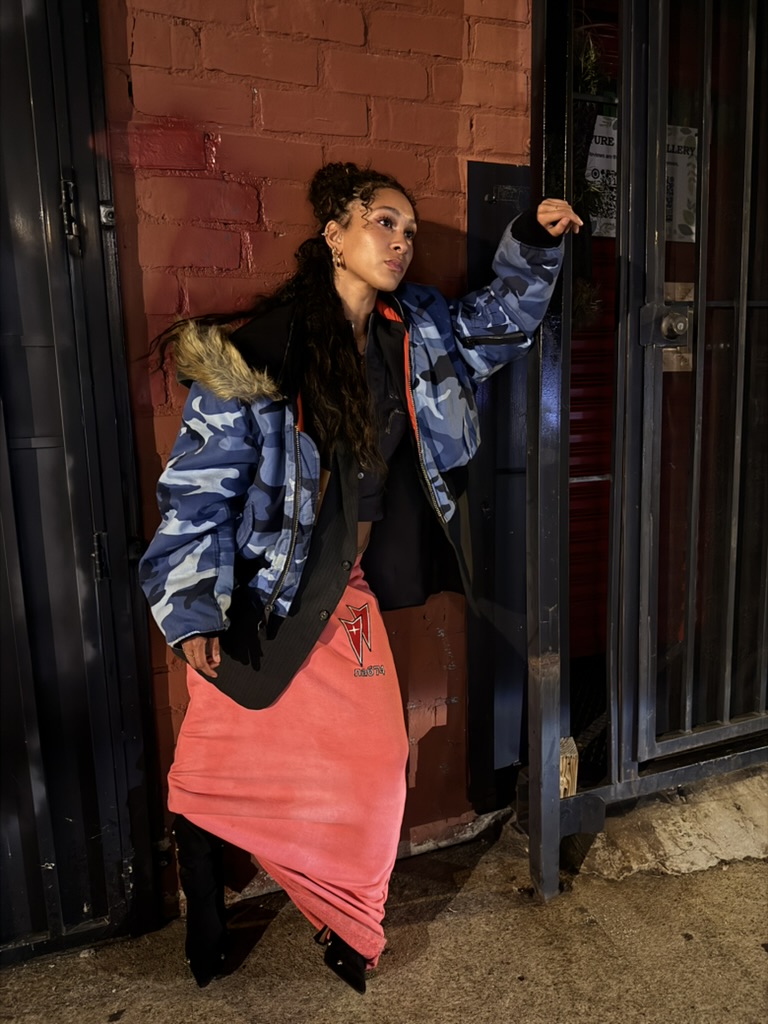 Maria Williams leaning against a red brick wall at night in a blue camo bomber with fur hood and coral maxi skirt for Lab 74