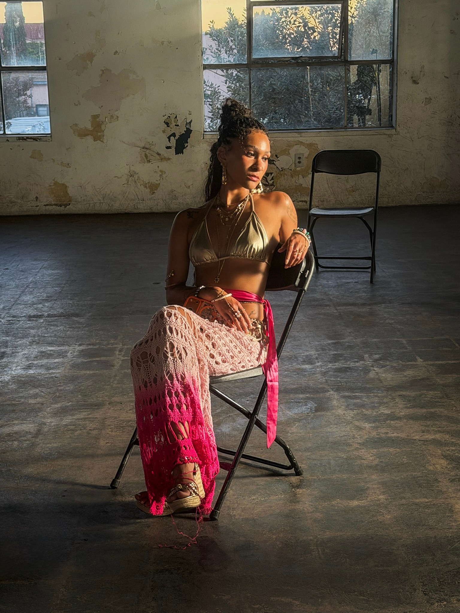 Maria Williams seated on a folding chair in a sunlit warehouse in a gold bikini top and pink ombré crochet pants on the Destin Conrad “Nervous” set