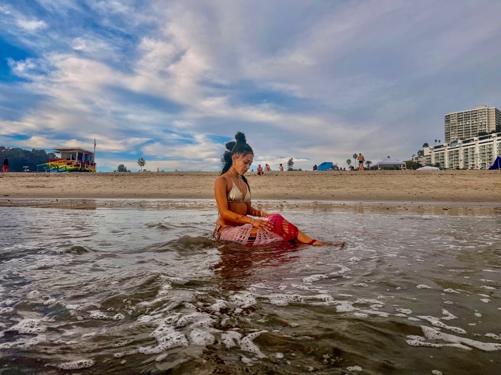 Maria Williams seated in the shallows looking down, lifeguard tower behind