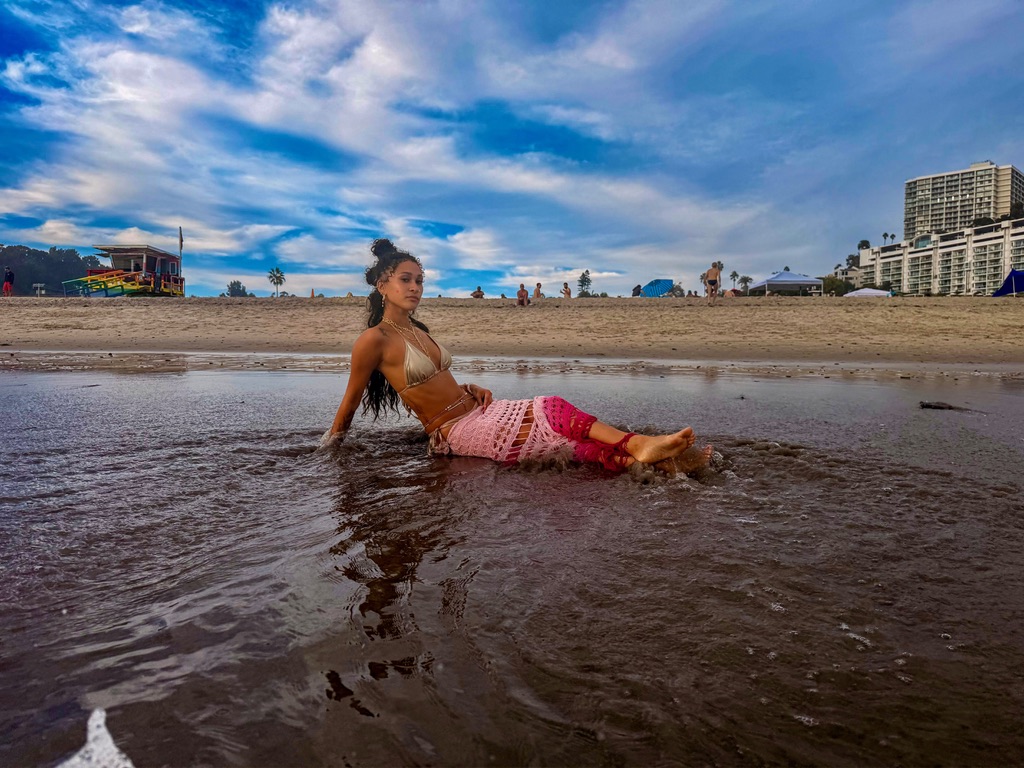 Maria Williams reclining in the shoreline water in a gold bikini and pink crochet skirt, her reflection beneath her