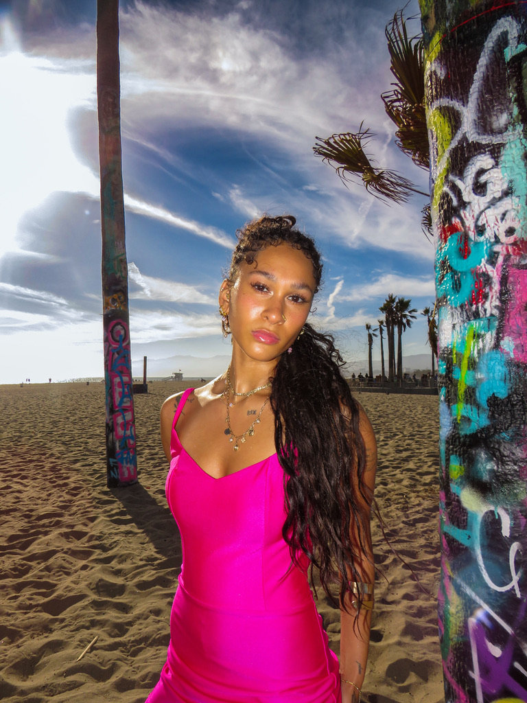 Maria Williams in a hot pink slip dress between graffiti-painted palm trunks on Venice Beach