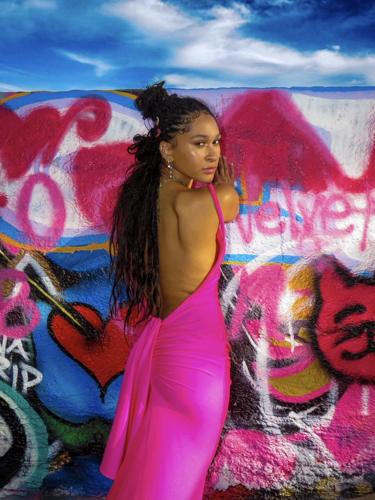 Maria Williams in a backless pink gown looking over her shoulder against a pink graffiti wall