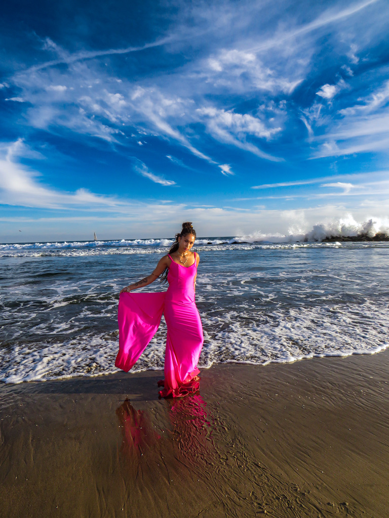 Maria Williams holding out the train of a hot pink gown at the shoreline as waves break behind her