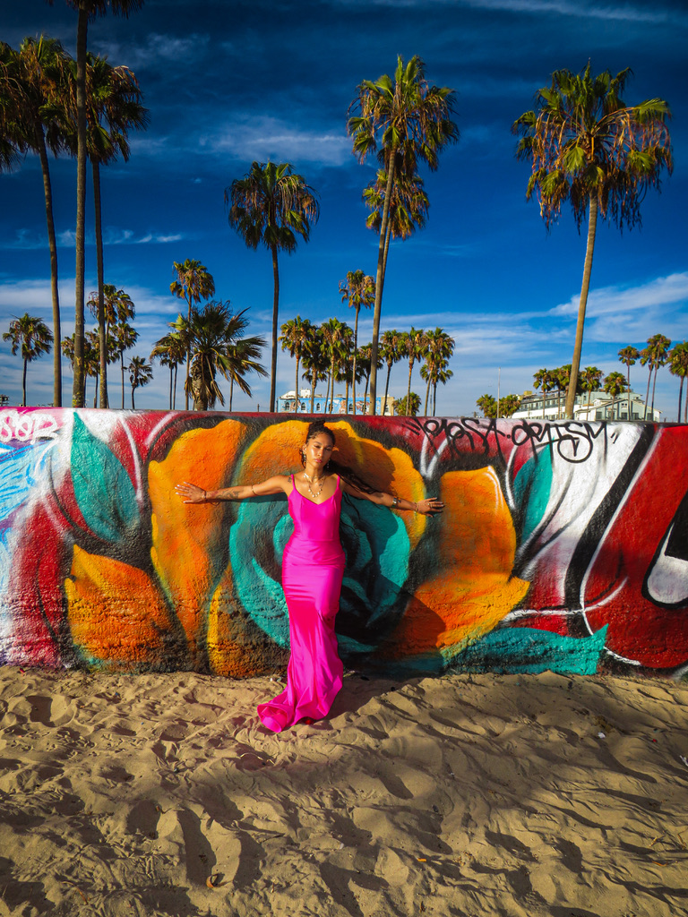Maria Williams in a hot pink gown with arms outstretched against a giant painted flower mural, palm trees above