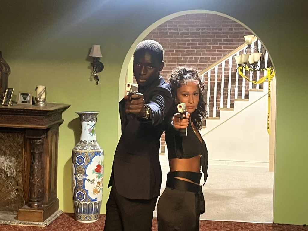 Two actors standing back to back aiming prop pistols beneath an arched doorway on the Writerboy film set