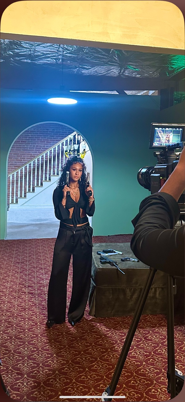 Behind the scenes on Writerboy: actress in black holding two prop pistols under an arch as a cinema camera films